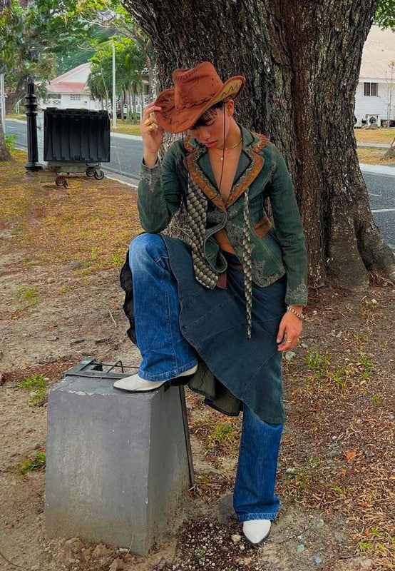 Person sitting on a concrete block outdoors, wearing a green jacket, blue jeans, and a brown hat.